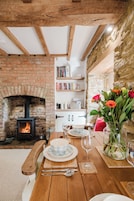 A cozy dining area with a brick fireplace, wooden beams, and a table set for two with plates, cutlery, and a vase of flowers.