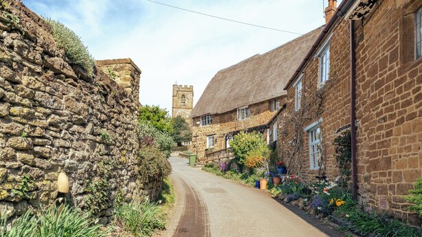 A narrow cobblestone street flanked by stone buildings with thatched roofs, a church tower in the distance, and a garden with colorful flowers. - Sweetheart Cottage-Cosy 2BD in Cotswolds (Banbury)