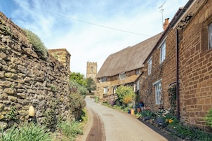 A narrow cobblestone street flanked by stone buildings with thatched roofs, a church tower in the distance, and a garden with colorful flowers.
