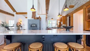 A modern kitchen with a central island, wooden bar stools, pendant lighting, and a stone fireplace. - Birch Beach Lake House-Hot tub/Sauna, Lake Access (Goderich)