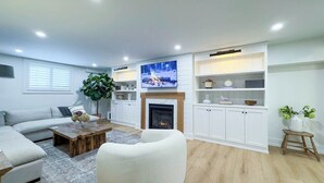 A modern living room with a fireplace, white furniture, and a large TV mounted above it. - Birch Beach Lake House-Hot tub/Sauna, Lake Access (Goderich)