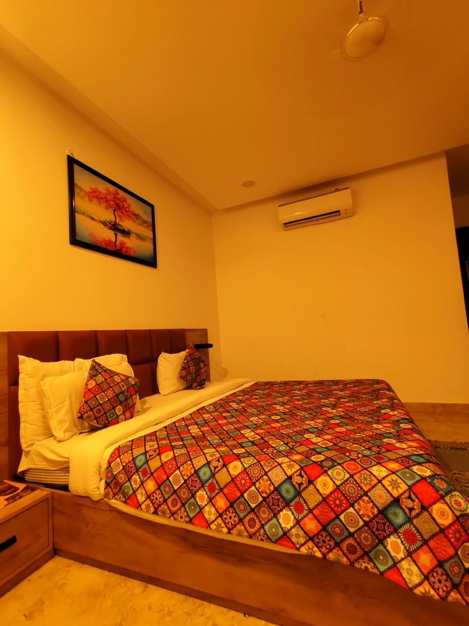 A hotel room with a bed, a nightstand, a wall-mounted air conditioner, and a framed picture.