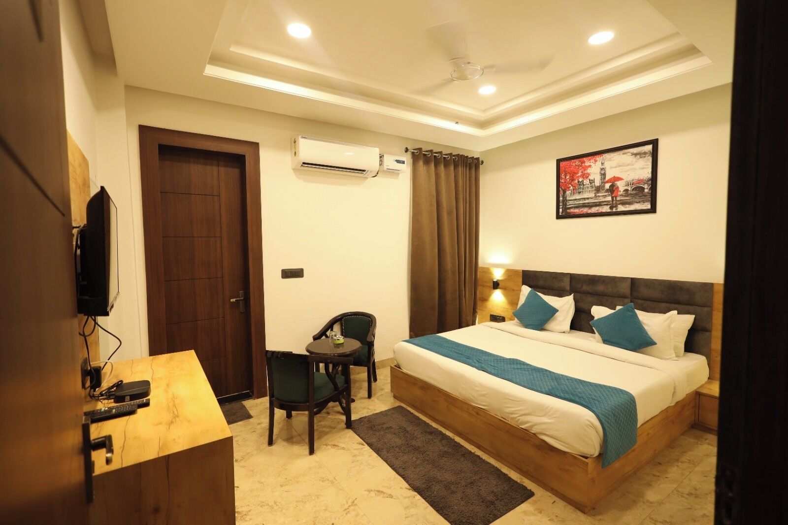 A hotel room with a bed, a desk, a chair, an air conditioning unit, and a painting on the wall.