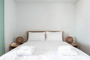 A neatly made bed with white linens, two bedside tables with wicker lamps, and a plain white wall.