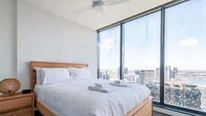 A modern hotel room with a large bed, wooden headboard, and a city view through large windows. - City Views Apt - FREE Parking , WIFI, Gym, + Pool! (Melbourne)