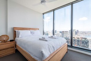 A modern hotel room with a large bed, wooden headboard, and a city view through large windows. - City Views Apt - FREE Parking , WIFI, Gym, + Pool! (Melbourne)