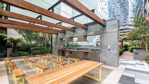 An outdoor seating area with wooden benches and tables, a modern kitchen, and a view of tall buildings. - City Views Apt - FREE Parking , WIFI, Gym, + Pool! (Melbourne)