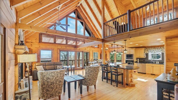 Interior espacioso de cabaña con techo alto, vigas de madera, comedor, cocina y zona de estar acogedora con sillas estampadas.
