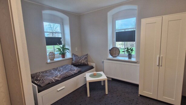 Ein kleines Zimmer mit einem Bett, einem Heizkörper, einem Fenster, einer Pflanze und einem Kleiderschrank.