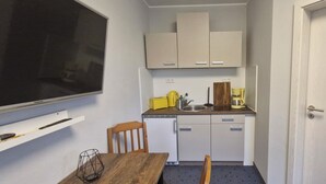 Eine kleine Küche mit einem weißen Kühlschrank, einer Spüle und einer Kaffeemaschine. Ein Holztisch mit einem Stuhl und ein an der Wand befestigtes Fernsehgerät sind ebenfalls vorhanden.