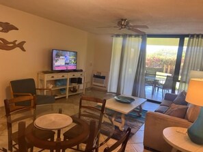 A living room with a television, a dining table, chairs, a sofa, and a balcony with curtains.