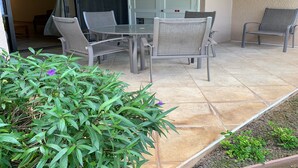 A patio with a glass table and chairs, a sliding door, and a small garden area. - Kamaole Sands #4-110 Ground Floor, Full A/C, Great Location, Fitness Center! (Kihei)
