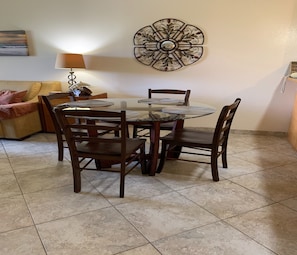 A dining area with a round glass table, four wooden chairs, a decorative wall piece, a lamp, and a sofa.