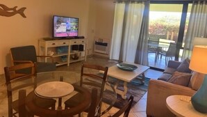 A living room with a television, a dining table, chairs, a sofa, and a balcony with curtains. - Kamaole Sands #4-110 Ground Floor, Full A/C, Great Location, Fitness Center! (Kihei)