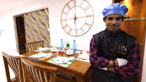 A dining area with a wooden table set for two, a large wall clock, and a door with a wooden frame. - 3 Bedroom Apartment ( Gagal Home) (Mumbai)