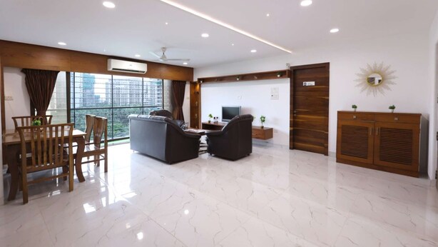 A modern living room with a dining area, a flat-screen TV, and a large window with a city view. - 3 Bedroom Apartment ( Gagal Home) (Mumbai)