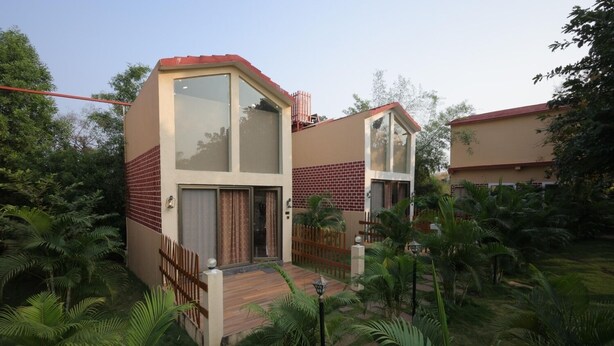 Una casa moderna de dos pisos con techo rojo, ventanas amplias y un jardín con plantas tropicales.