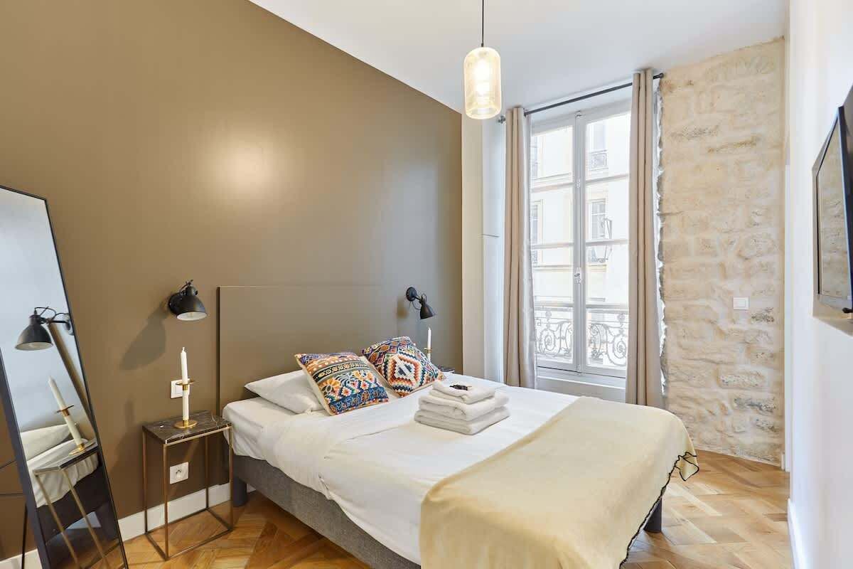 Une chambre à coucher avec un lit, une table de chevet, une lampe et une fenêtre donnant sur des immeubles.