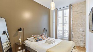 A bedroom with a bed, bedside table, lamp, and a window with a view of buildings.