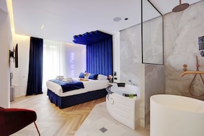 A modern hotel room with a large bed, a white vanity, a bathtub, and a large window with blue curtains.
