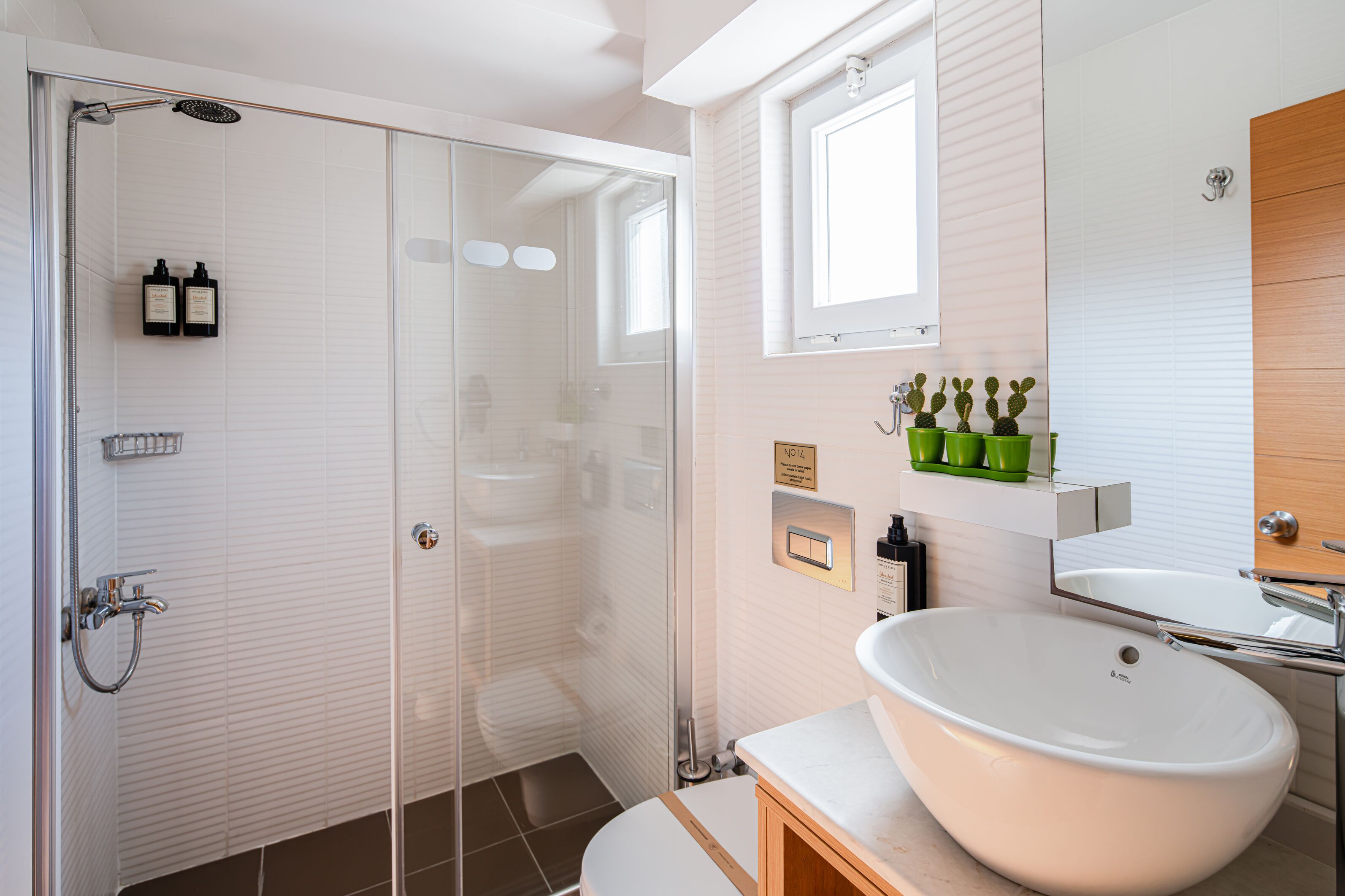 A bathroom with a glass shower enclosure, a white sink, and a window above the mirror.