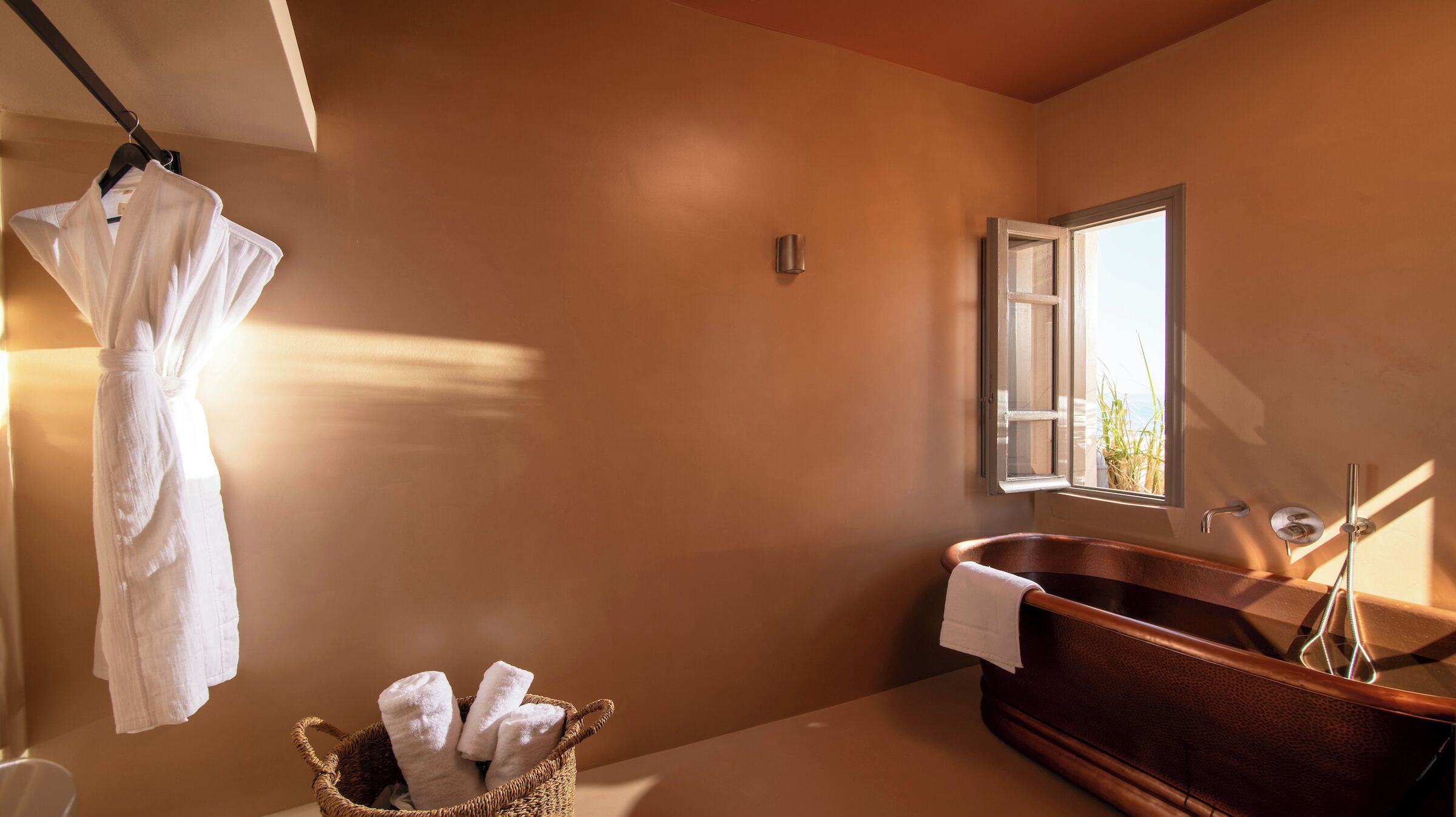A bathroom with a wooden bathtub, a hanging robe, and a basket of towels.