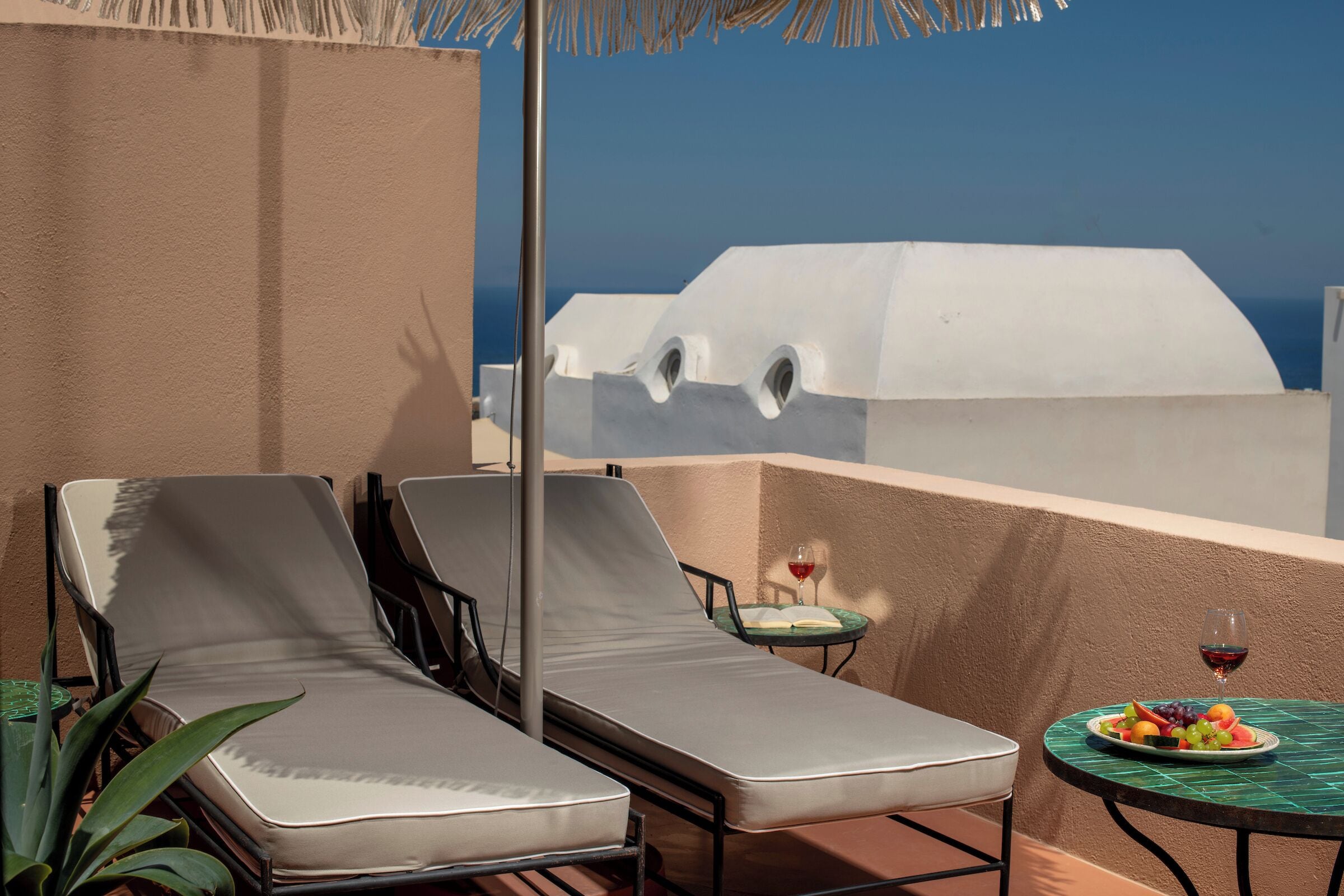 A balcony with two lounge chairs, a small table with a plate of fruit and a glass of red wine, and a view of a white building against a clear blue sky.