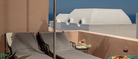 A balcony with two lounge chairs, a small table with a plate of fruit and a glass of red wine, and a view of a white building against a clear blue sky.
