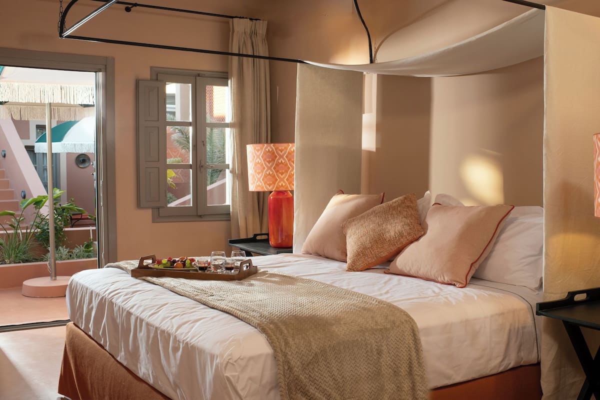 Een slaapkamer met een bed, nachtkastjes, een schaal met fruit en glazen, een raam met gordijnen, en uitzicht op een trap en groen buiten.