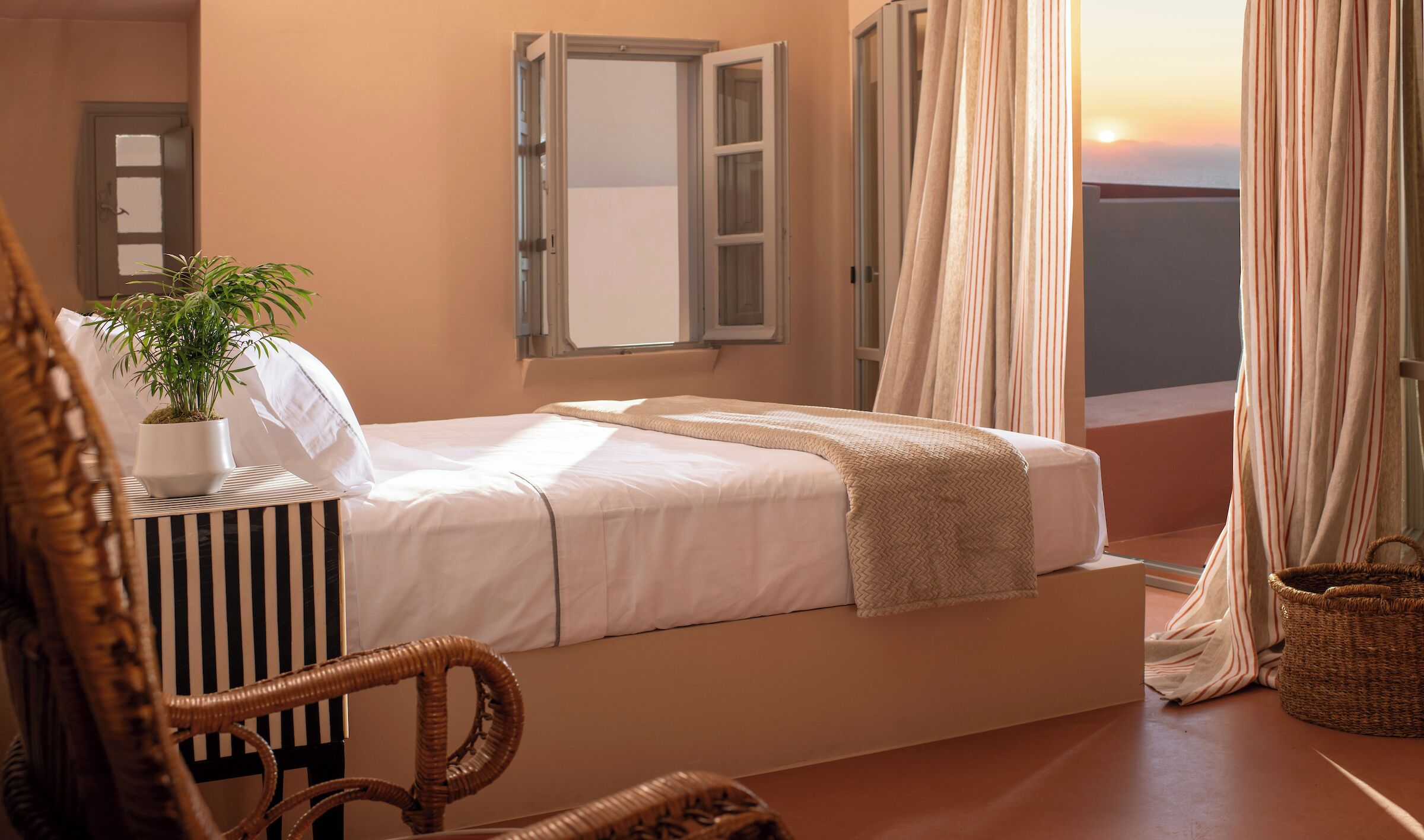 A bedroom with a bed, a potted plant, a wicker chair, and a view of the sunset through the window.