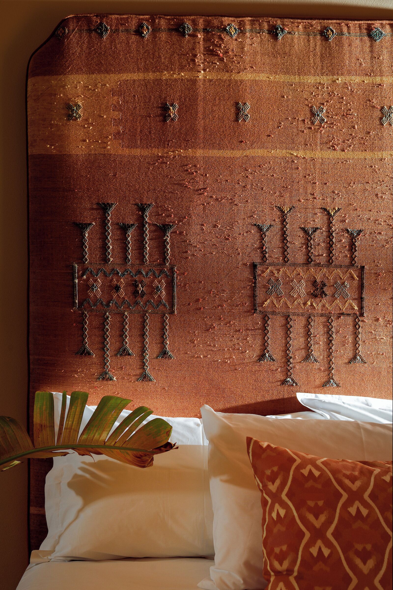 A bed with white linens and a patterned pillow, a large decorative headboard with intricate designs, and a plant in the foreground.