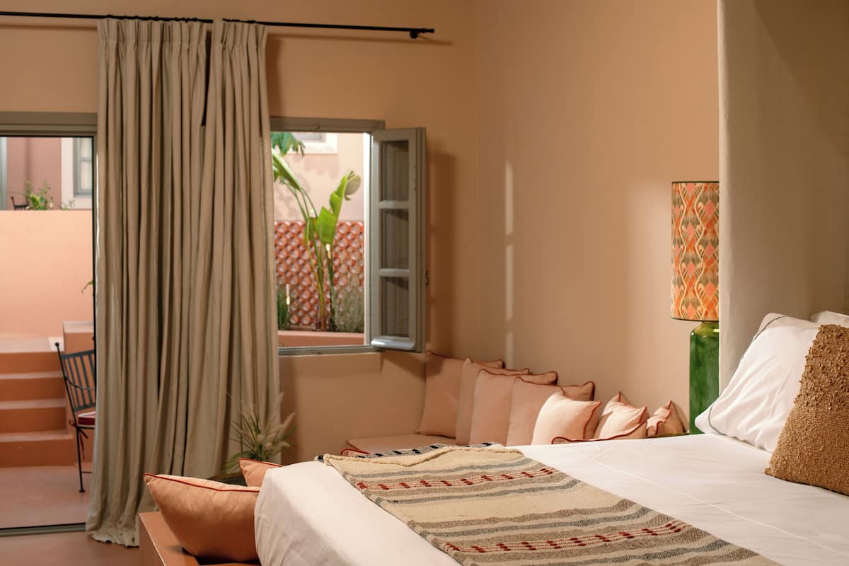 Een slaapkamer met een bed, kussens, een raam met gordijnen, een stoel en een lamp.