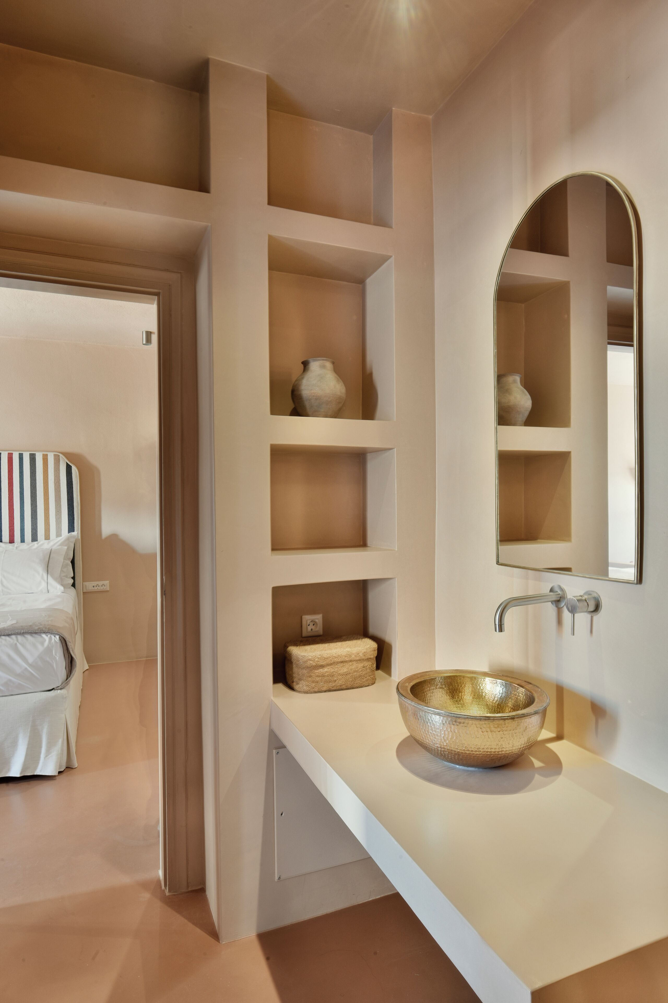 A modern bathroom with a white sink, a gold bowl, and a mirror. There are shelves with decorative items and a striped headboard in the background.