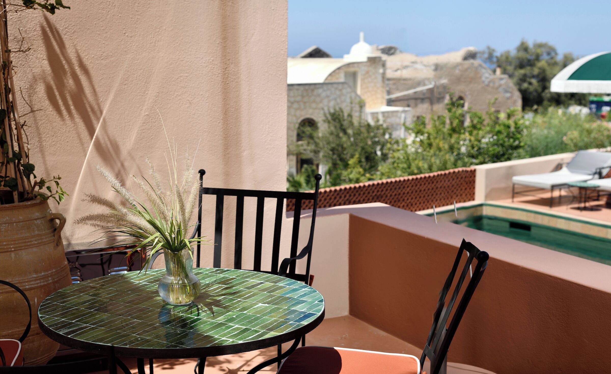 A round glass table with a vase of plants, a chair, and a view of a pool and buildings in the background.
