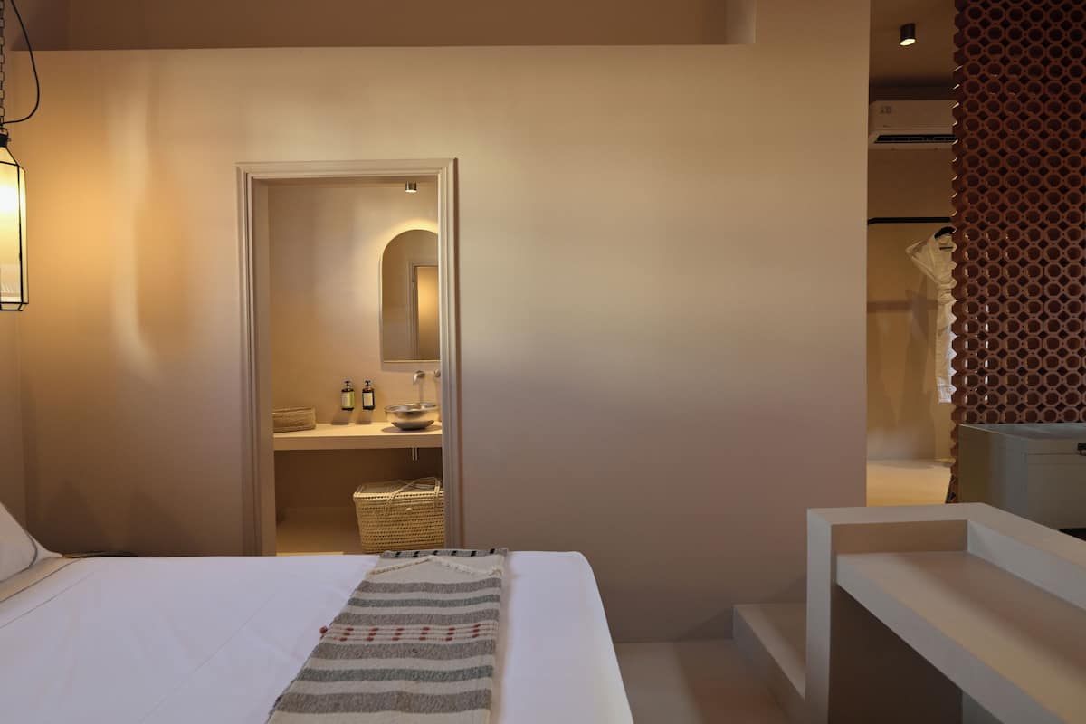 Een slaapkamer met een bed, een nachtkastje, een hanglamp, een spiegel en uitzicht op een badkamer met een wastafel en toiletartikelen.