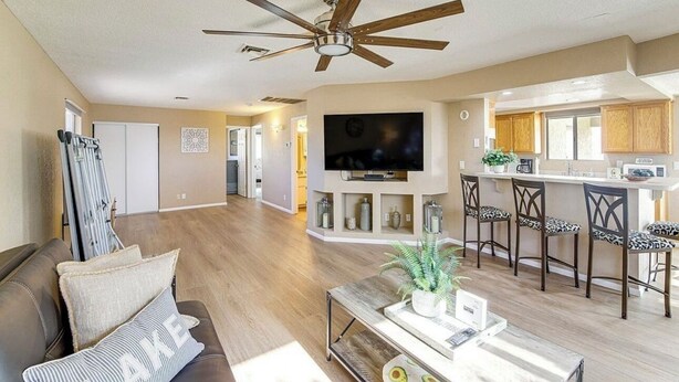 A spacious living room with a ceiling fan, a TV, a sofa, and a kitchen area with wooden cabinets and bar stools.