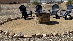 An outdoor seating area with Adirondack chairs and a stone fire pit, surrounded by a gravel area and a chain-link fence.