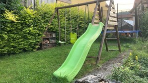 Un columpio de madera con un tobogán verde en un patio trasero con cerco de madera y follaje.