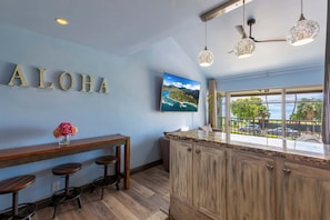 A kitchen with a wooden bar, a 'ALOHA' sign, a painting of a landscape, and a view of the outdoors through a window.