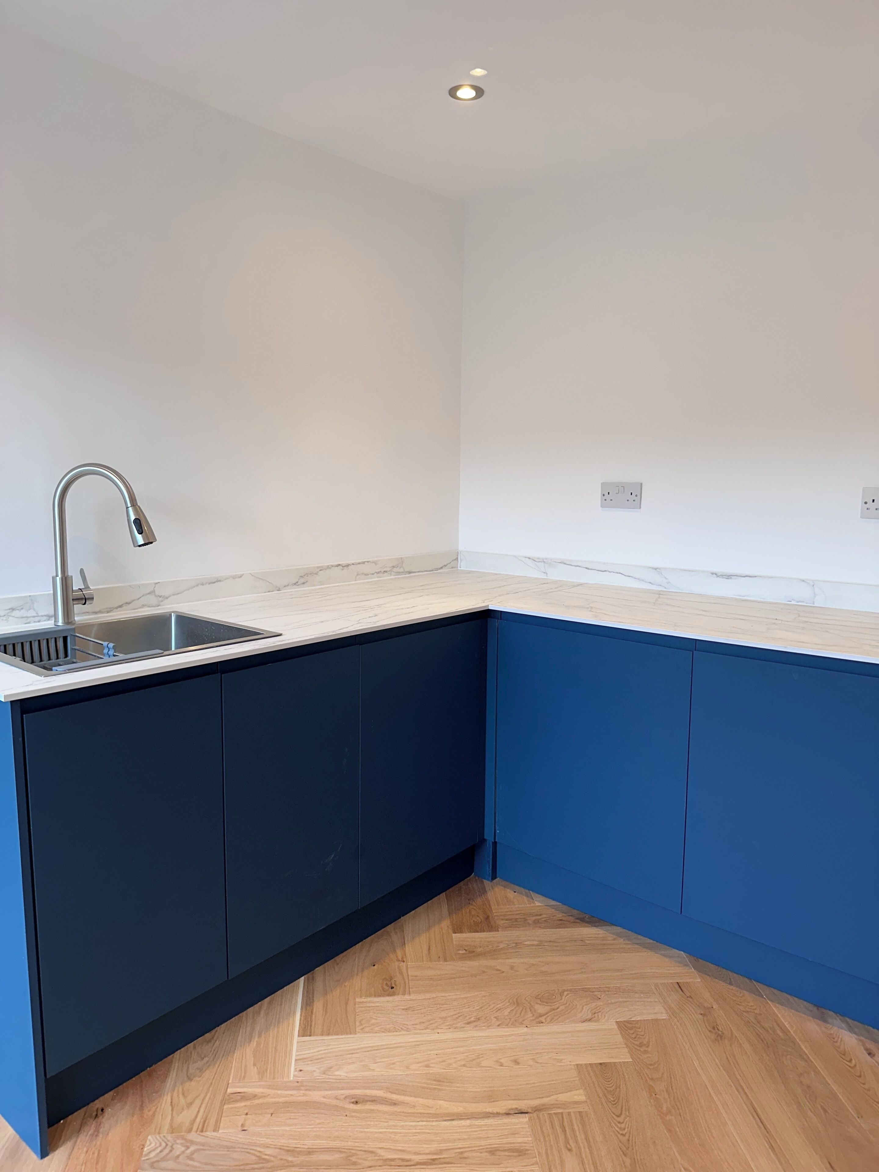Une cuisine dotée d’un évier encastré, de meubles de rangement bleu foncé, d’un plan de travail en marbre et d’un parquet en bois disposé en chevrons.
