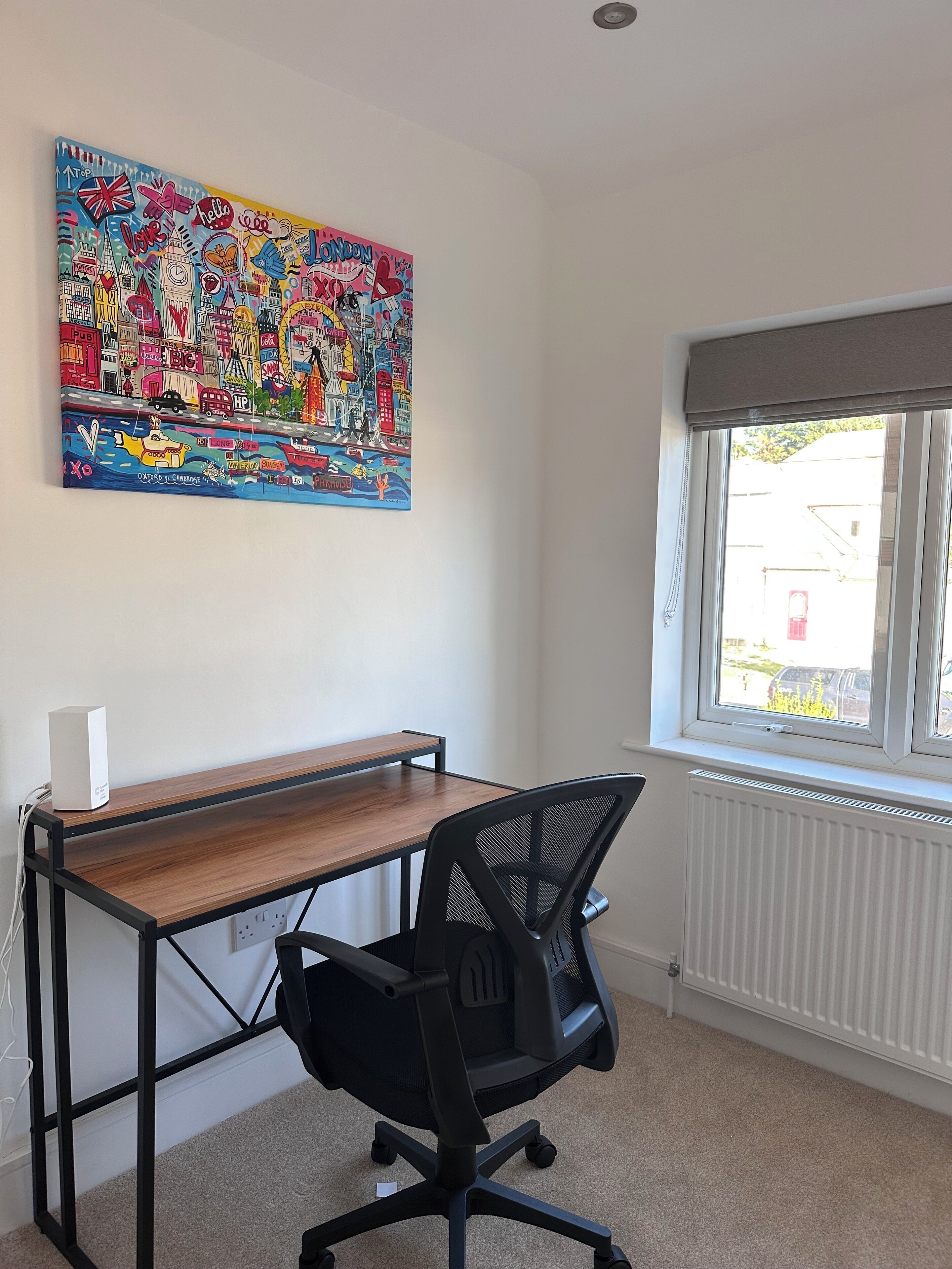 A room with a wooden desk, a black office chair, a colorful wall art depicting London landmarks, and a window with a view of the outside.