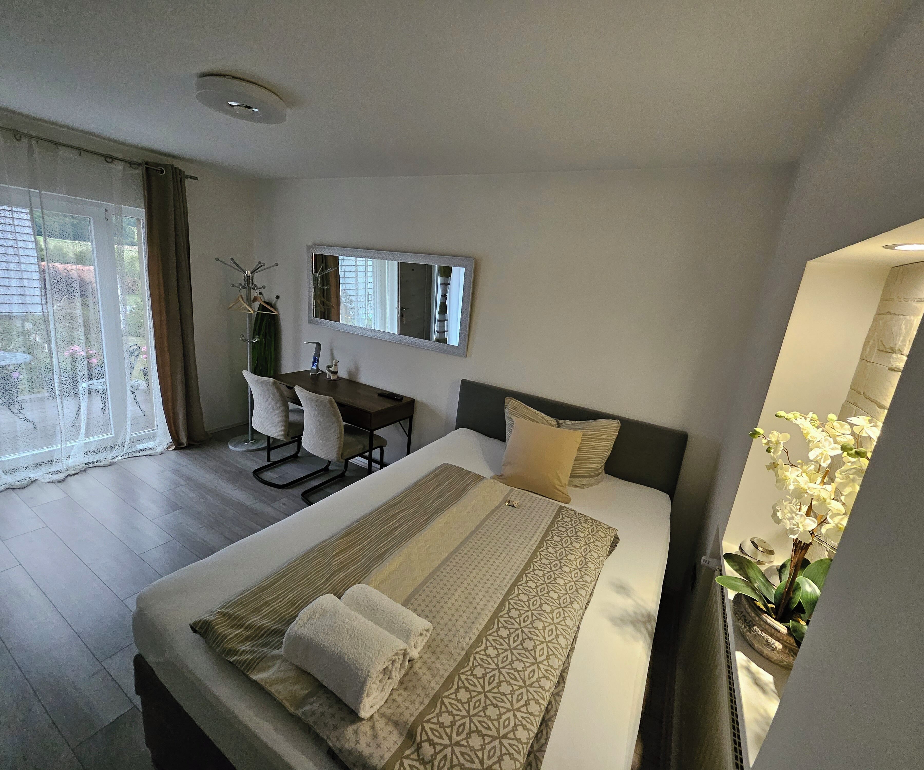Une chambre d’hôtel avec un lit, un bureau, une chaise, un miroir et une plante en pot.
