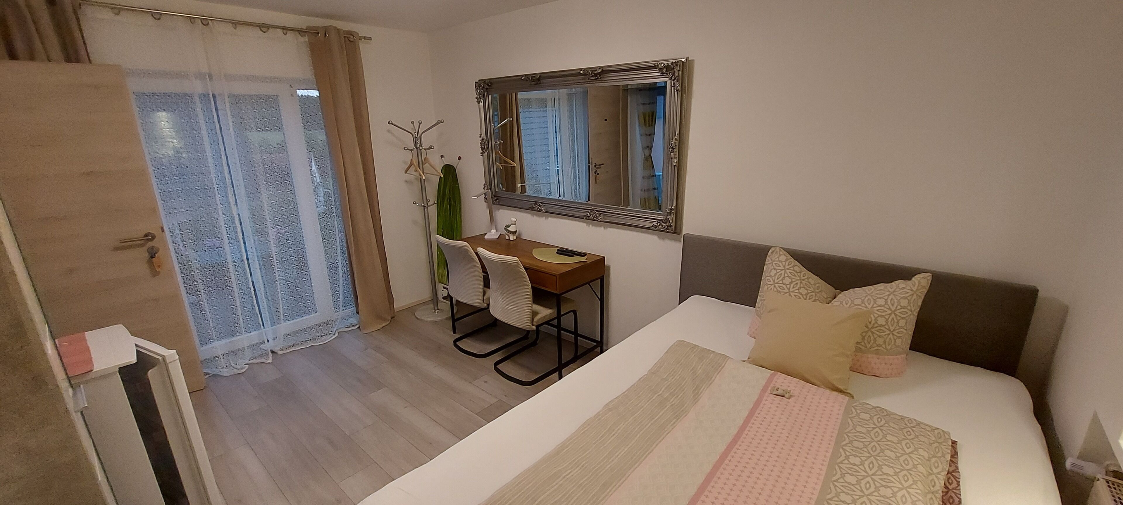 A hotel room with a bed, a desk with a chair, a mirror, and a window with curtains.