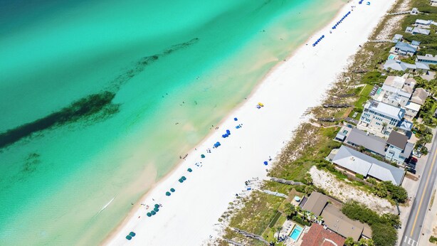 Vue aérienne d’une plage aux eaux turquoise et limpides, bordée d’un rivage sablonneux et d’une rangée de maisons dotées d’une piscine.