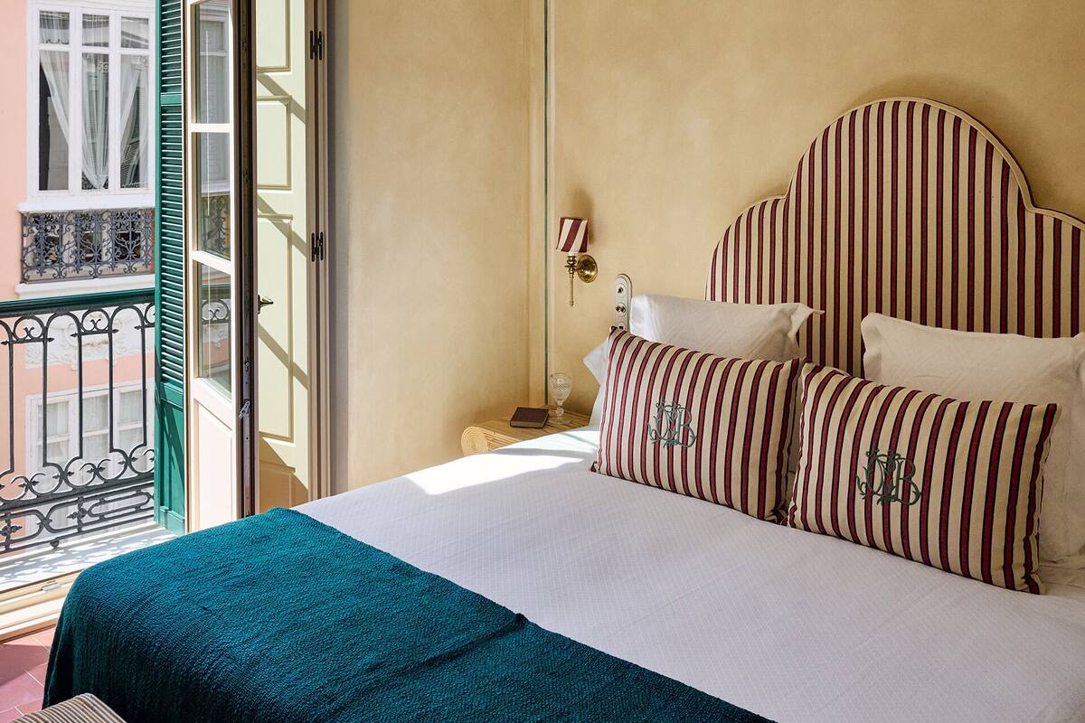 A hotel room with a bed, striped pillows, a nightstand, a lamp, and a view of the outside balcony.