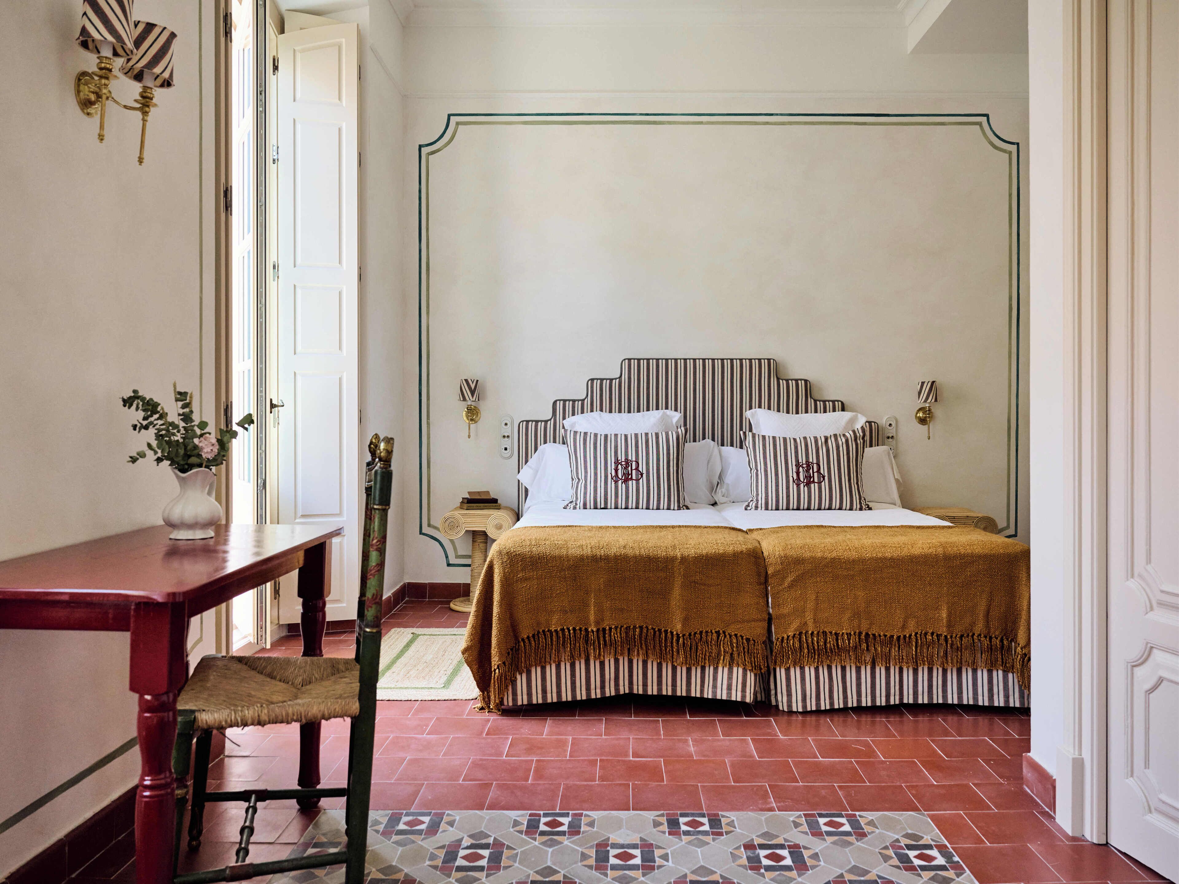 Un dormitorio con cama, mesitas de noche, una silla, un escritorio y un florero con flores.