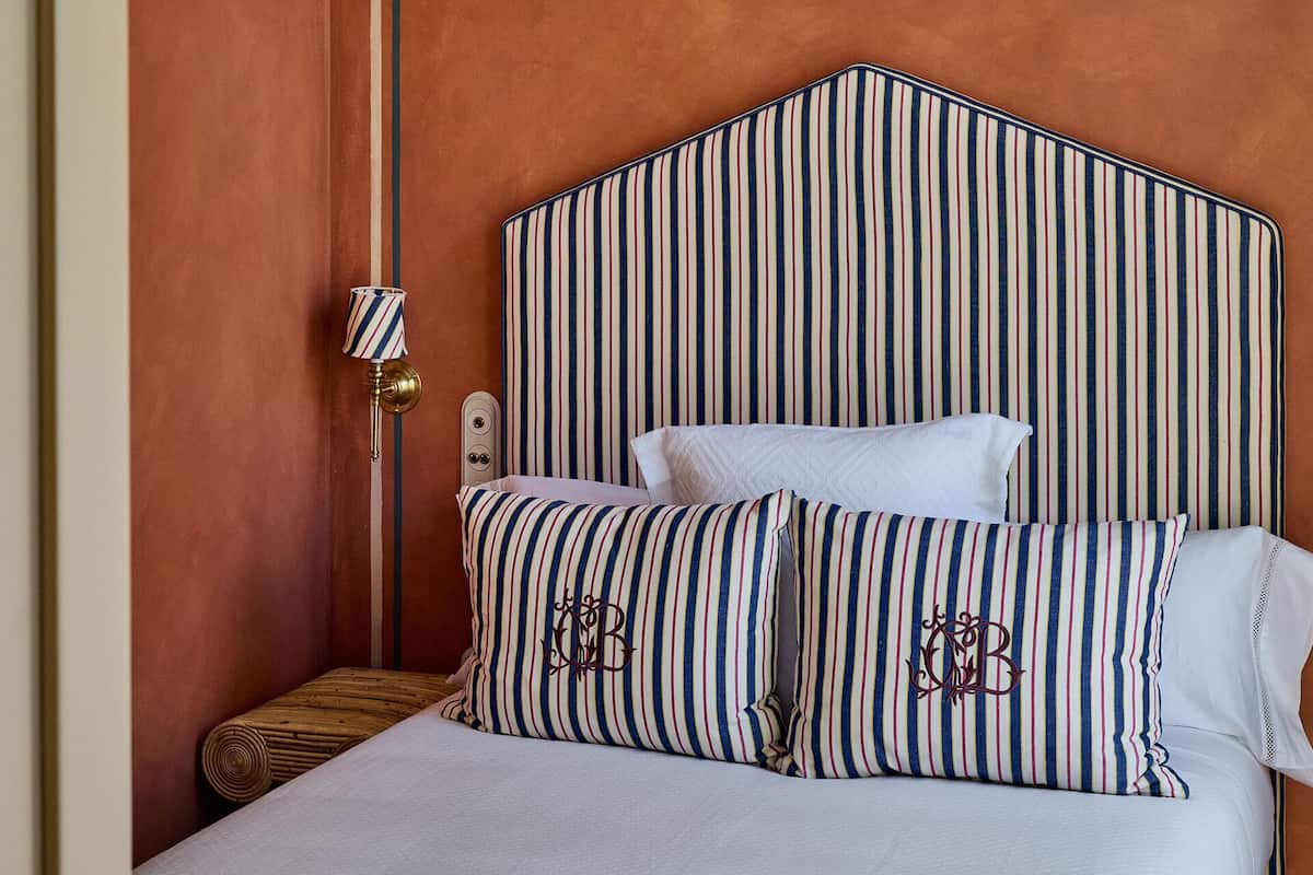 A hotel room with a striped headboard, a bedside lamp, and two pillows with embroidered initials.