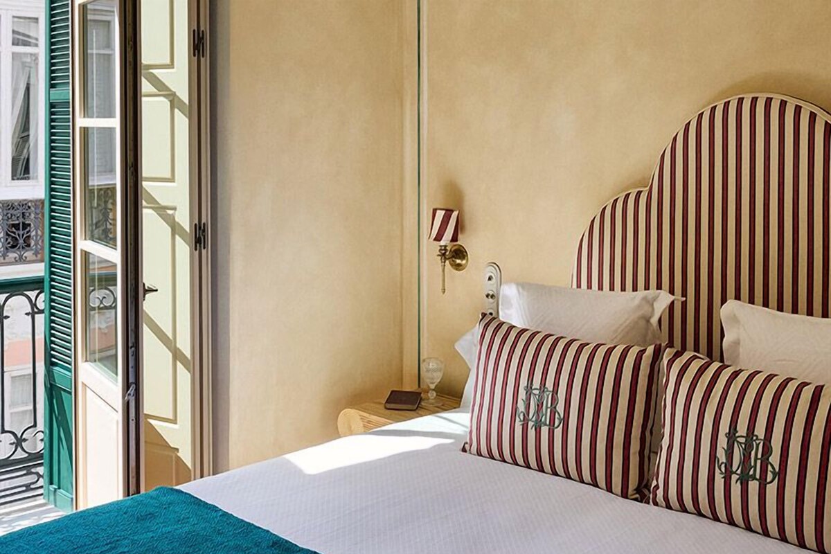 A hotel room with a large bed, striped pillows, a bedside table with a lamp, and a view of the outside.