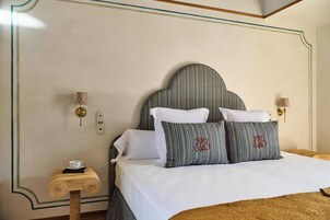 Una cama bien hecha con cabecera a rayas, ropa de cama blanca y dos almohadas con iniciales bordadas. Una mesita de madera con una taza y un libro están al lado de la cama.