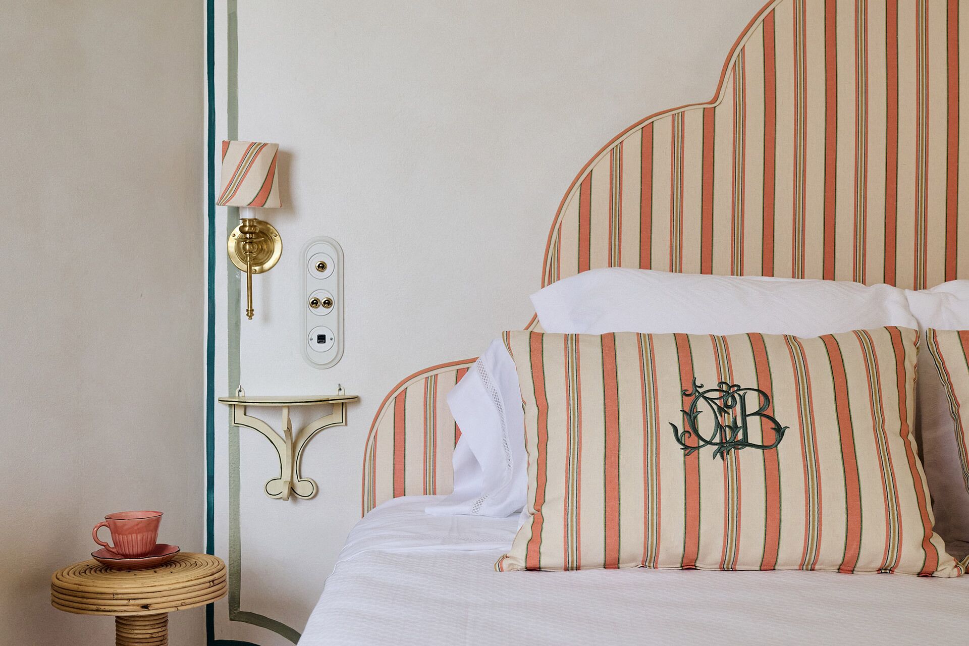 A hotel room with a bed featuring striped bedding, a bedside table with a cup and saucer, and a wall-mounted light fixture.
